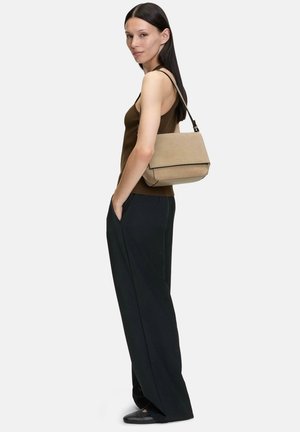 Sac à épaule en suédine beige au design structuré, de forme rectangulaire et avec des bordures noires. Le modèle le présente avec un débardeur marron et un pantalon noir.