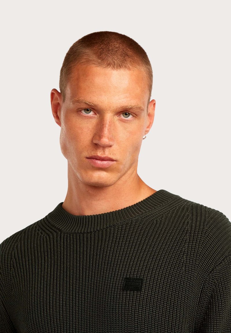 Pull en tricot vert foncé avec une texture côtelée. Il présente un col rond et une petite étiquette logo sur la poitrine.