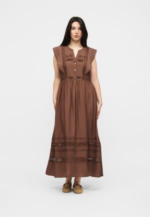 Femme portant une robe midi marron sans manches avec des détails en dentelle, des boutons sur le corsage et des chaussures plates tissées, debout devant un fond uni.