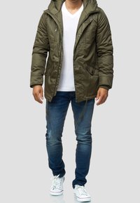 Oliwkowa, ocieplana parka z kapturem i zapięciem na zamek błyskawiczny, z dwoma bocznymi kieszeniami i ściągaczami na dolnym brzegu, zestawiona z wypłowiałymi niebieskimi dżinsami i białymi trampkami.