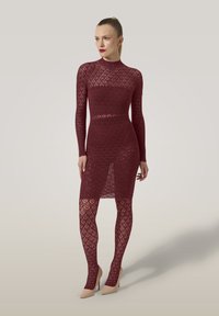 Kvinde iført en tætsiddende, langærmet, burgunderrød blondekjole med matchende mønstrede tights og beige højhælede sko, stående foran en ensfarvet baggrund.
