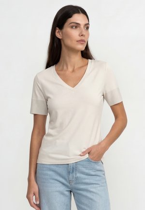 Žena s dlhými tmavými vlasmi, ktorá má na sebe krémové tričko s výstrihom do V a krátkymi rukávmi a svetlomodré džínsy, s jednou rukou v vrecku, pozerá sa doprava.