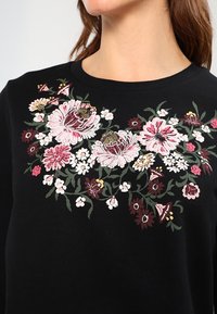 Sweat-shirt noir avec une broderie florale colorée dans des nuances de rose, blanc et bordeaux, avec des détails de feuillage vert sur la poitrine.