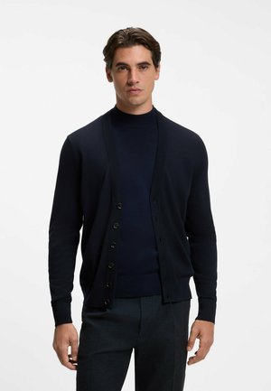Mann mit dunklen Haaren, der eine schwarze, knöpfbare Strickjacke über einem schwarzen Rollkragenpullover und dunklen Hosen trägt, stehend vor einem weißen Hintergrund.