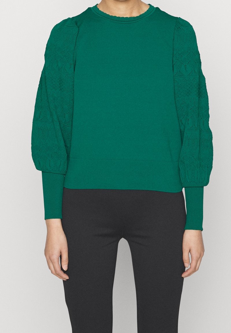 Pull vert avec manches en dentelle texturée et bouffantes, poignets côtelés et encolure festonnée. Tissu tricoté doux, cintré à la taille.