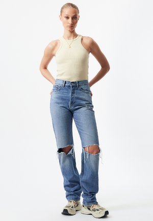 Ribbed beige tank top paired with light blue high-waisted jeans featuring distressed knee rips. Model wears beige and black sneakers.
