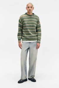 Πλεκτό πουλόβερ με πράσινες ρίγες και μπεζ λεπτομέρειες, συνδυασμένο με φωτεινά μπλε παντελόνια φαρδιάς γραμμής και μαύρα παπούτσια. Το μοντέλο στέκεται μπροστά σε απλό φόντο.