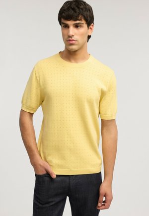 Uomo che indossa una maglia gialla a maniche corte in maglia con piccoli fori, abbinata a jeans scuri, in piedi davanti a uno sfondo semplice.