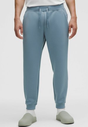 Jasnoniebieskie spodnie dresowe z elastycznym pasem i sznurkiem, wyposażone w boczne kieszenie i dopasowane ściągacze przy kostkach. Noszone z miękkimi zielonymi chodakami.
