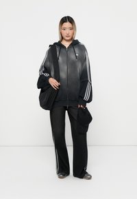 Svart zip-up hoodie med vita ränder, i kombination med mörka vida byxor och en svart keps. Outfiten har sportiga designelement och en avslappnad look.