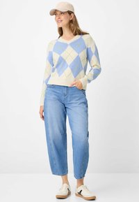 Maglioncino leggero con scollo a V, caratterizzato da un motivo a rombi blu, crema e verde. Abbinato a jeans blu a gamba larga e sneakers.