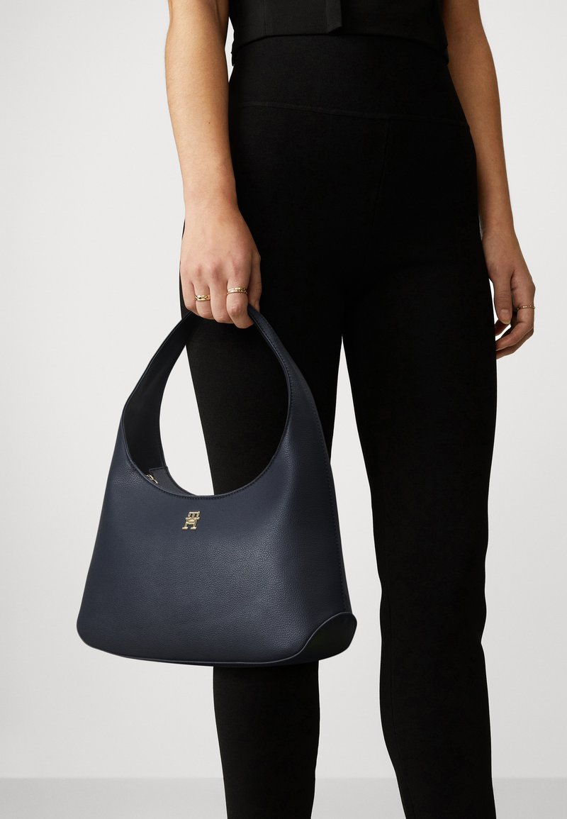 Navyblaue Hobo-Tasche aus strukturiertem Leder, mit einem gebogenen Griff und einem goldenen Logo-Akzent auf der Vorderseite.