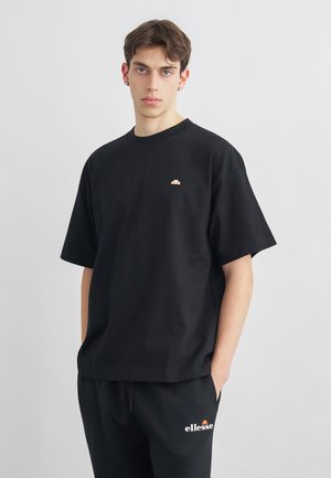 Jonge man met kort bruin haar, gekleed in een zwart ellesse T-shirt en broek, staand tegen een lichtgrijze achtergrond, handen in de zakken.