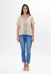 Camisa de lino beige de manga corta y cuello en V, con rayas verticales y detalles de trabajo de calado, combinada con jeans de mezclilla azules.