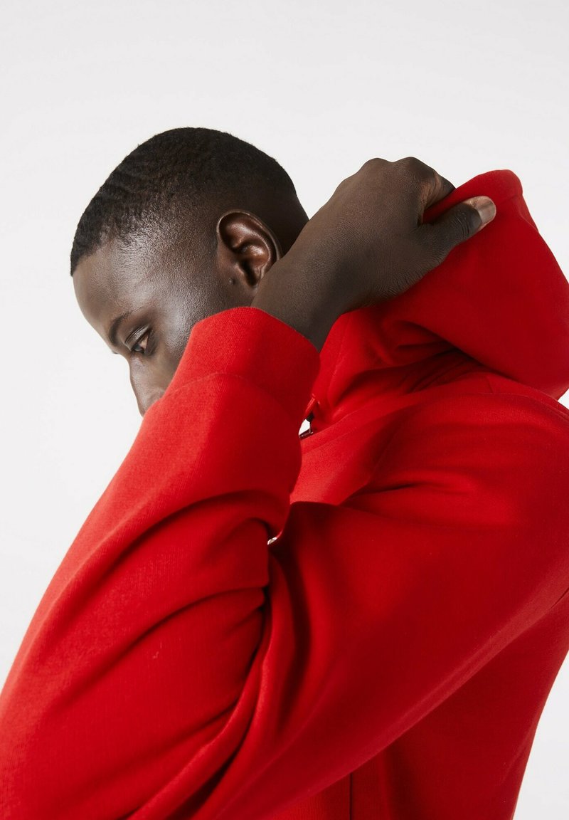 Rote Kapuzenjacke mit einer strukturierten Oberfläche, die eng anliegt und eine verstellbare Kordelkapuze hat. Die Ärmels sind eng anliegend und mit Rippbündchen versehen.