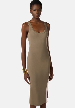 Donna che indossa un abito midi beige senza maniche con scollo a barca e spacco laterale, accessoriato con un ampio bracciale rigido in oro al polso sinistro.