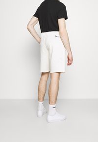 Lätta herrshorts i krämfärg med elastisk midja och svart Nike-logotyp, kombinerade med en svart t-shirt samt vita skor och strumpor.