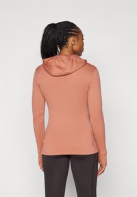 Top de manga longa com capuz em tecido rosa suave, apresentando um design ajustado com buracos para os polegares, finalizado com uma textura suave e detalhes de costura subtis.