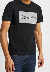 Camiseta de algodón negra con un parche rectangular gris que lleva el texto "Calvin Klein" en negro. Mangas cortas y escote redondo.