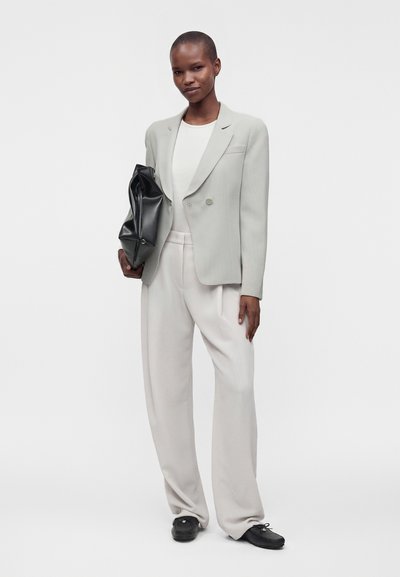 Personne portant un costume gris clair et un haut blanc tient une grande pochette noire, debout devant un fond blanc uni.