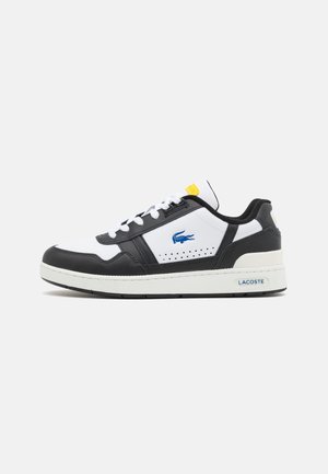 Hvid og sort lavtop sneaker med blåt Lacoste krokodille-logo, hvide snørebånd og gul mærkat på tungen, sidesål branding på ensfarvet baggrund.