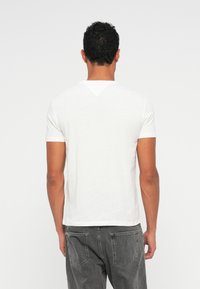 Λευκό βαμβακερό T-shirt, κοντά μανίκια, στρογγυλή λαιμόκοψη, απλή σχεδίαση. Φορεμένο με γκρι τζιν παντελόνι, από πίσω.