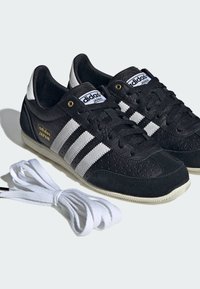Czarne buty sportowe Adidas Japan z białymi paskami, białymi akcentami na pięcie, złotym logo i dodatkowymi białymi sznurowadłami obok nich, na jasnym tle.