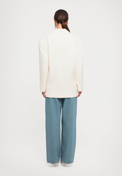 Vrouw met bruin haar in een lage paardenstaart, gekleed in een oversized witte blazer, wijdvallende blauwe jeans en beige hakken, staande met de rug naar de kijker gekeerd.