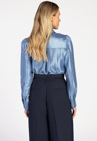 Satijnen blauwe blouse met pofmouwen en een achterpassant detail, gecombineerd met navy hoge taille broek met zijzakken.
