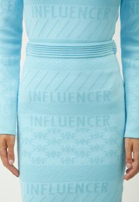 Hellblau gestricktes Kleid mit dem Wort "INFLUENCER" in verschiedenen Farbtönen. Das Design umfasst ein strukturiertes Band um die Taille.