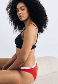 Fekete vezeték nélküli melltartó alacsony szabású piros bugyival, amelyen fehér Calvin Klein derékpánt található. Göndör hajú modell egy semleges háttérrel ül.