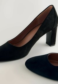 Black suede high-heeled pumps with a pointed toe and a solid block heel. Interior features a smooth brown lining. Subtle texture across material.