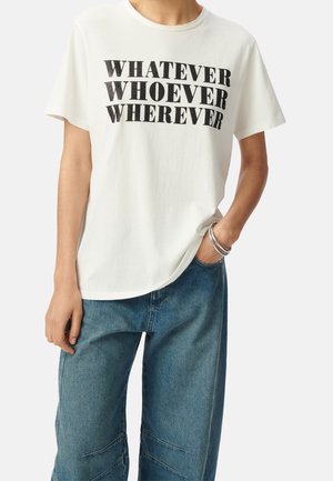 Osoba nosząca białą koszulkę z czarnym napisem "WHATEVER WHOEVER WHEREVER" i niebieskie dżinsy, jedna ręka w kieszeni z srebrnymi bransoletkami.