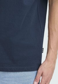 Camiseta de algodón de color azul marino con mangas cortas, etiqueta lateral y textura sutil. Las mangas y el dobladillo tienen costuras limpias. Se combina con denim claro.