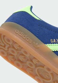 Niebieski zamszowy sneaker Adidas Gazelle z neonowo zielonymi paskami i przezroczystą gumową podeszwą z wytłoczonym logo Adidas.