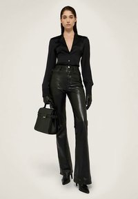 Černá na míru šitá košile s dlouhými rukávy a zapínáním na knoflíky, doplněná černými koženými kalhotami s vysokým pasem a rozšířenými nohavicemi. Černá kabelka a rukavice dokončují vzhled.