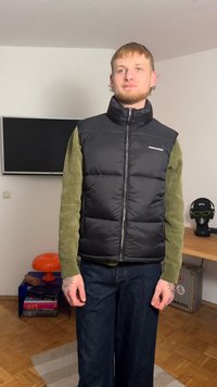 Colete preto acolchoado com gola alta e fecho de correr frontal, usado por cima de um suéter de malha verde. Combinado com jeans escuros de perna larga.