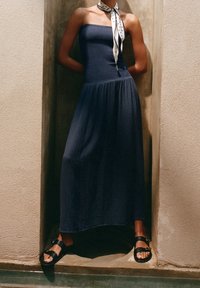 Robe bustier bleu marine en tissu texturé, jupe fluide ; associée à des sandales noires et un foulard blanc à pois autour du cou.