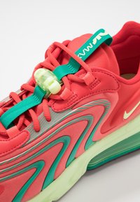 Röd och teal sportskor med vågiga teal-accenter, teal dragflik, vit luftkuddesula och grön snörlås på röda snören.