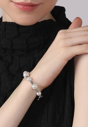 Perlenarmband mit fünf weißen Perlen und silbernen Akzenten, mit einem Verschluss. Getragen an einer Hand mit einem strukturierten schwarzen Oberteil im Hintergrund.