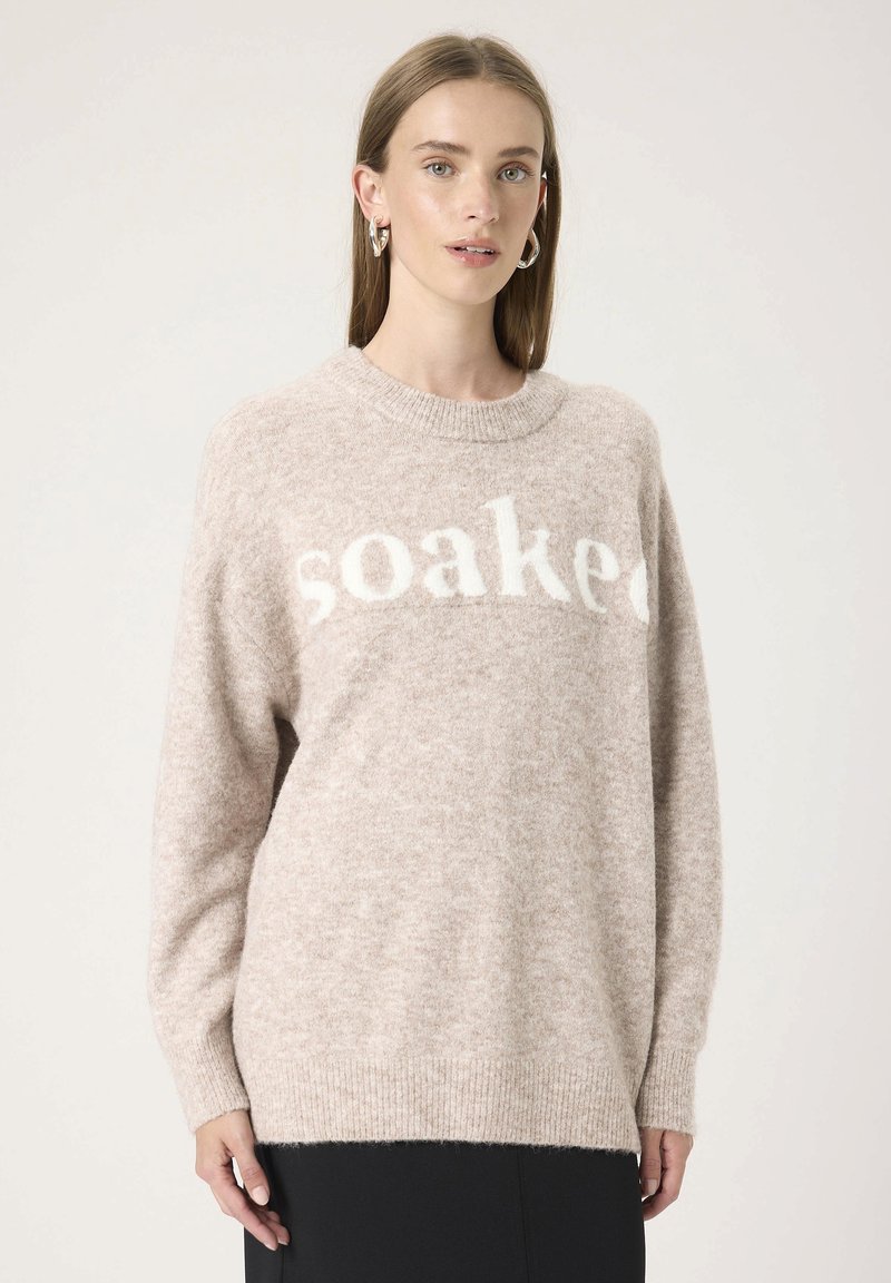 Femme portant un pull en laine beige avec le texte "soaked", des boucles d'oreilles en argent, se tenant devant un fond clair uni.