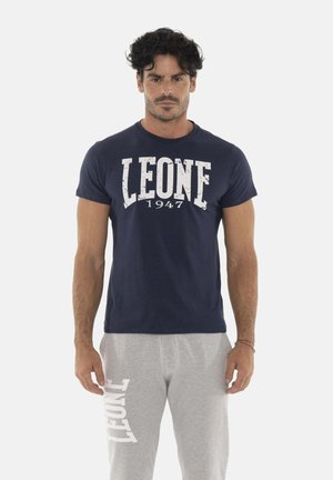 Man staat met het gezicht naar voren, gekleed in een marineblauw "LEONE 1947" T-shirt en lichtgrijze trainingsbroek met "LEONE" gedrukt op het linkerbeen.