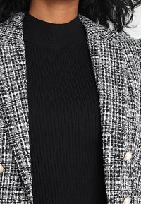 Blazer en tweed noir et blanc avec épaules structurées et grands boutons crème sur un haut noir côtelé à col montant.