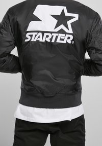 Giubbotto bomber nero con texture lucida, logo bianco oversize "STARTER" e design a stella sul retro, polsini e orlo a costine.