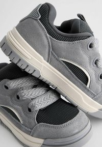 Grå sneakers med en overdel af ruskind og mesh, teksturerede snørebånd, gummisål og en diskret logo-detail på siden.