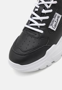 Sneaker en faux cuir noir avec des détails perforés, lacets blancs et une épaisse semelle blanche ; présente un patch logo sur le côté.