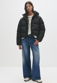 Jonge vrouw in een zwarte pufferjack, witte blouse, blauwe jeans met wijde pijpen, witte sneakers en zilveren kettingen tegen een effen achtergrond.