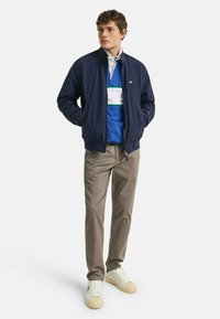 Chaqueta bomber azul marino sobre una polo azul con rayas blancas y verdes, combinada con pantalones beige y zapatillas blancas con acentos crema.