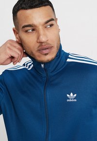 Modrá zipová sportovní bunda se bílými trojpruhy na ramenou a malým bílým logem Adidas na hrudi. Hladká textilie.