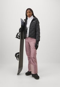 Schwarze steppte Jacke, weißer Rollkragenpullover, rosa Skihosen. Modell hält ein Snowboard, trägt schwarze Handschuhe und Schuhe. Schlichter, klarer Hintergrund.